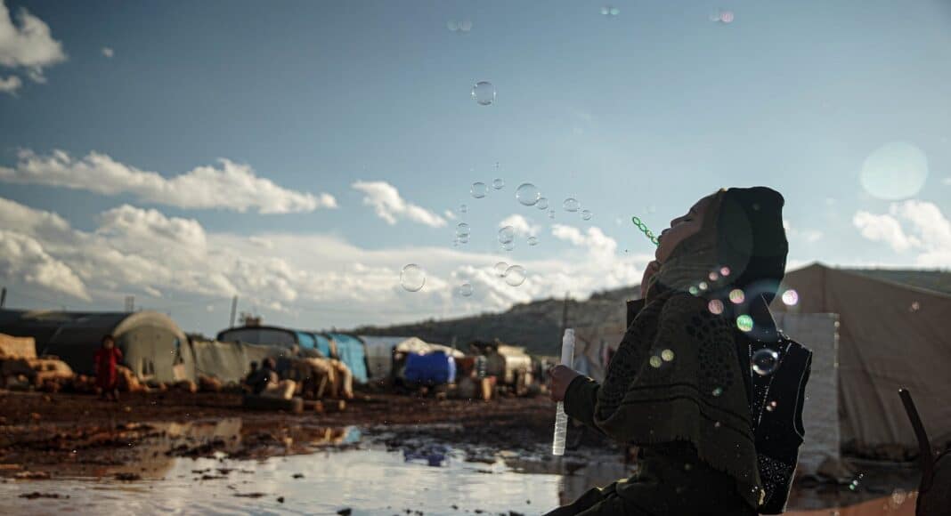 hijabi woman kneeling by puddle in refugee camp blowing bubbles