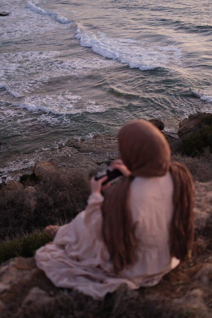 woman in brown hijab sitting on the cliff near the ocean